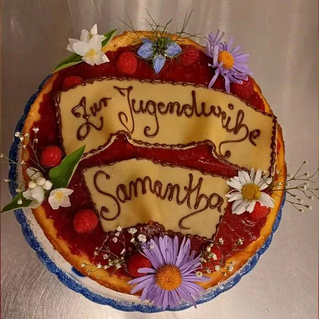 Käsekuchen mit Himbeeren, Blüten und beschriftetem Marzipanbanner zur Jugendweihe vom Café am Markt in Jena